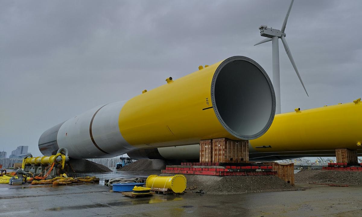 Large yellow steel segments on supports in a port, a wind turbine behind them